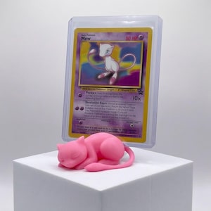 Op de afbeelding: Een roze Mew-beeldje in slaaphouding staat op een witte kubus, met een Mew Pokémon-kaart in een beschermhoes erachter. De kaart toont een paarse en witte Mew-illustratie met tekst. Het beeldje is een gladde sculptuur.