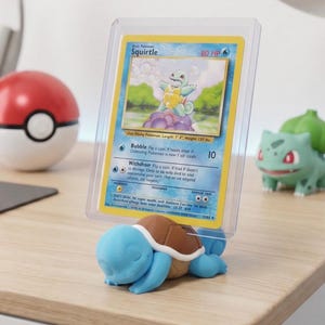 May include: A blue and brown Squirtle figure holding a Pokémon trading card in a protective case. The card features a Squirtle illustration with the text "Squirtle" and "80 HP". A red and white Pokéball and a green Bulbasaur figure are in the background.