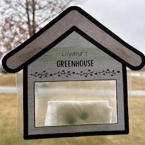 Puede incluir: Una casa de papel blanco con una ventana de plástico transparente, etiquetada como "Lilyana's GREENHOUSE". Una pequeña planta verde está creciendo dentro de la ventana.
