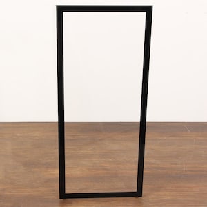 May include: A tall, rectangular, vertical frame with a smooth, solid black finish. The frame features clean, straight edges and a minimalist, open center design, providing a stark contrast against the surrounding environment.