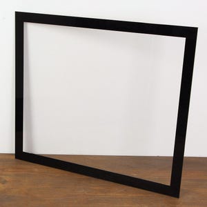 Könnte beinhalten: Ein schwarzer rechteckiger Rahmen, vor einem weißen Hintergrund, liegt auf einer Holzoberfläche. Das schlanke, minimalistische Design des Rahmens deutet auf eine moderne Ästhetik hin, die sich für die Präsentation von Kunstwerken oder Fotos eignet.