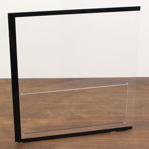 Op de afbeelding: Een heldere acryl vitrine met een zwart frame. De vitrine heeft een rechthoekige vorm en een horizontale plank. De achtergrond is een lichtbruin houten oppervlak.
