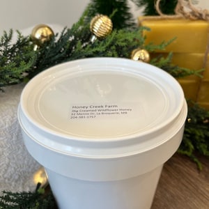 May include: A white plastic container with a white lid, labelled "Honey Creek Farm 3kg Creamed Wildflower Honey 32 Menno Dr, La Broquerie, MB 204-381-1757".