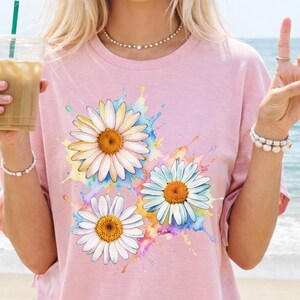 May include: A light pink t-shirt featuring a watercolor design of three daisies with white petals and orange centers. The daisies are surrounded by colorful paint splashes. The word "BLOSSOM" is printed at the bottom left.