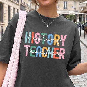 May include: A charcoal gray short-sleeve t-shirt featuring the text HISTORY TEACHER in a multi-colored, distressed block font. The word HISTORY is in blue, orange, and pink, while TEACHER is in pink, green, and orange.