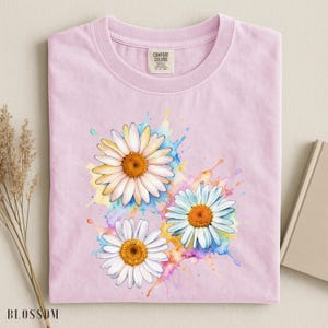 May include: A light pink t-shirt featuring three daisy flowers with white petals and orange centers. The daisies are set against a colorful watercolor splash design. The shirt has a Comfort Colors tag.