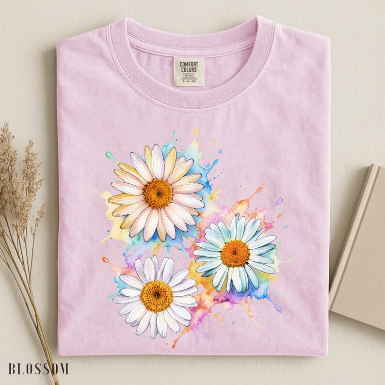 May include: A light pink t-shirt featuring three daisy flowers with white petals and orange centers. The daisies are set against a colorful watercolor splash design. The shirt has a Comfort Colors tag.