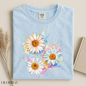 May include: Light blue t-shirt featuring a watercolor design of three daisies with white petals and orange centers. The daisies are set against colorful paint splatters. The shirt has a crew neck and short sleeves. The tag reads "COMFORT COLORS".
