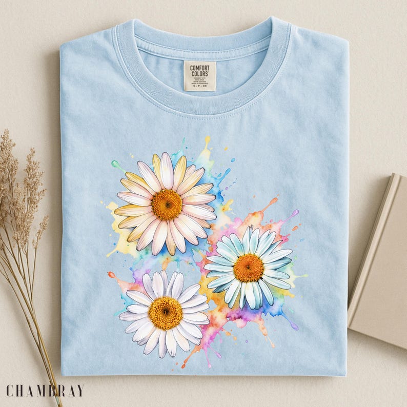 May include: Light blue t-shirt featuring a watercolor design of three daisies with white petals and orange centers. The daisies are set against colorful paint splatters. The shirt has a crew neck and short sleeves. The tag reads "COMFORT COLORS".