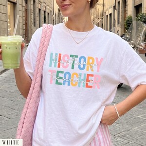 May include: A white short-sleeved t-shirt featuring the text "HISTORY TEACHER" printed in a multi-colored, distressed block font. The word "history" is in teal, orange, and green, while "teacher" is in pink, green, yellow, and orange.