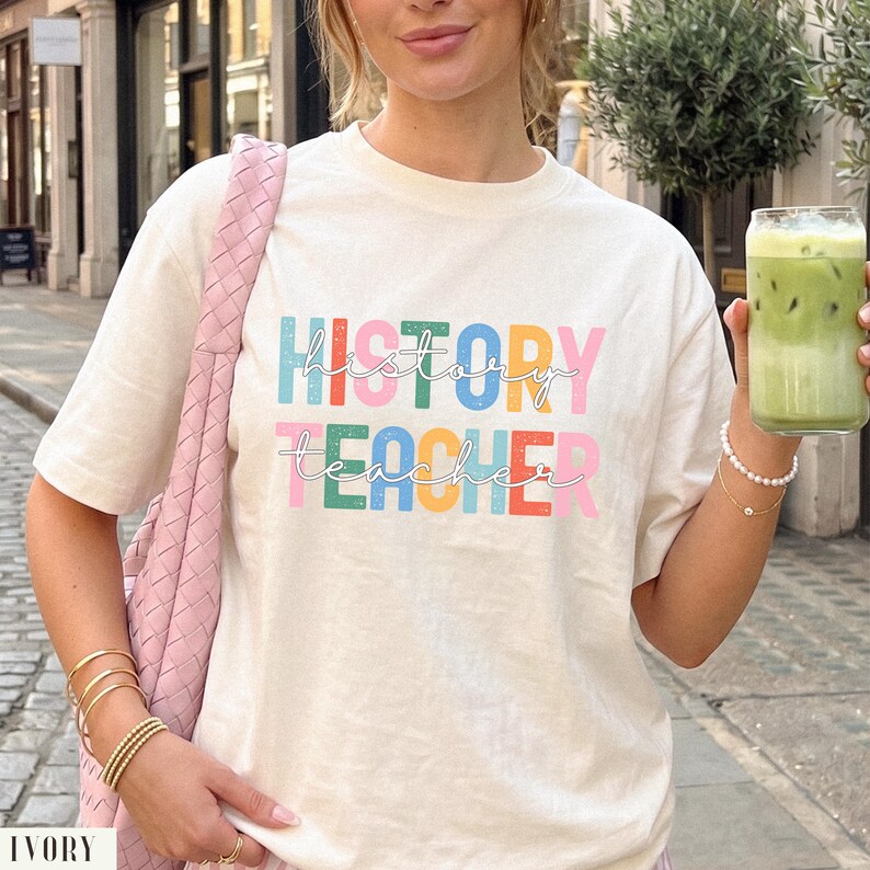 May include: An ivory short-sleeve t-shirt featuring the text "HISTORY TEACHER" printed in a multi-colored, block-letter style with a speckled texture. The word "history" is in blue, green, yellow, and pink, while "teacher" is in green, blue, yellow, red, and pink.