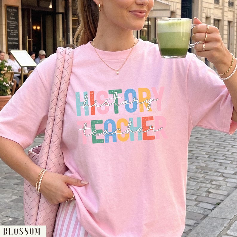 May include: A light pink short-sleeved t-shirt featuring the text "HISTORY TEACHER" printed in large, colorful block letters with a white cursive script overlay. The shirt is paired with a woven pink shoulder bag and a glass mug of green tea.