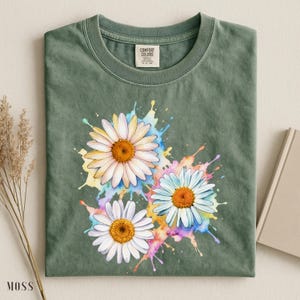 May include: Moss green t-shirt featuring a watercolor design of three daisies with white petals and orange centers. The daisies are set against colorful paint splashes. The shirt has a crew neck and a tag that says "Comfort Colors".