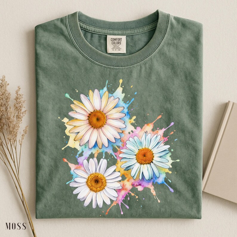 May include: Moss green t-shirt featuring a watercolor design of three daisies with white petals and orange centers. The daisies are set against colorful paint splashes. The shirt has a crew neck and a tag that says "Comfort Colors".