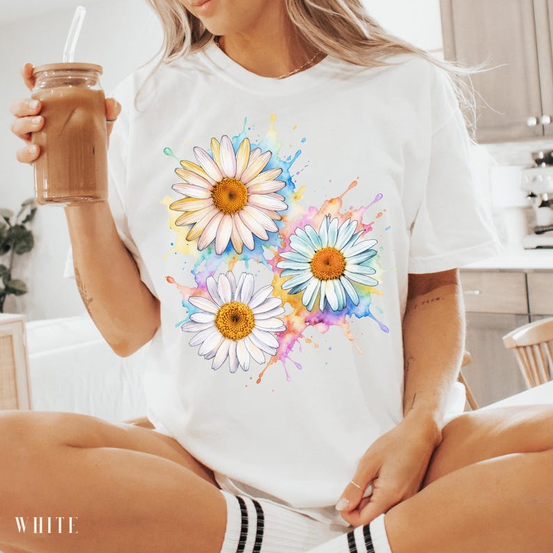 May include: White t-shirt featuring a watercolor design of three daisies with yellow and orange centers. The daisies are surrounded by colorful paint splatters. The word "WHITE" is printed at the bottom of the shirt.