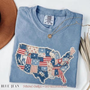 May include: A light blue Comfort Colors t-shirt with a printed design of the United States map. The map is made of patchwork fabric with red, white, and blue patterns, and is outlined with faux pearls. The text "BLUE JEAN" and "Printed design - not embroidered" are at the bottom.
