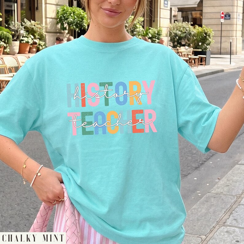May include: A light turquoise short-sleeve t-shirt featuring the text "history teacher" printed in a multi-colored block font. The word "history" is in light blue, orange, green, blue, yellow, and pink, while "teacher" is in pink, green, yellow, blue, and pink.