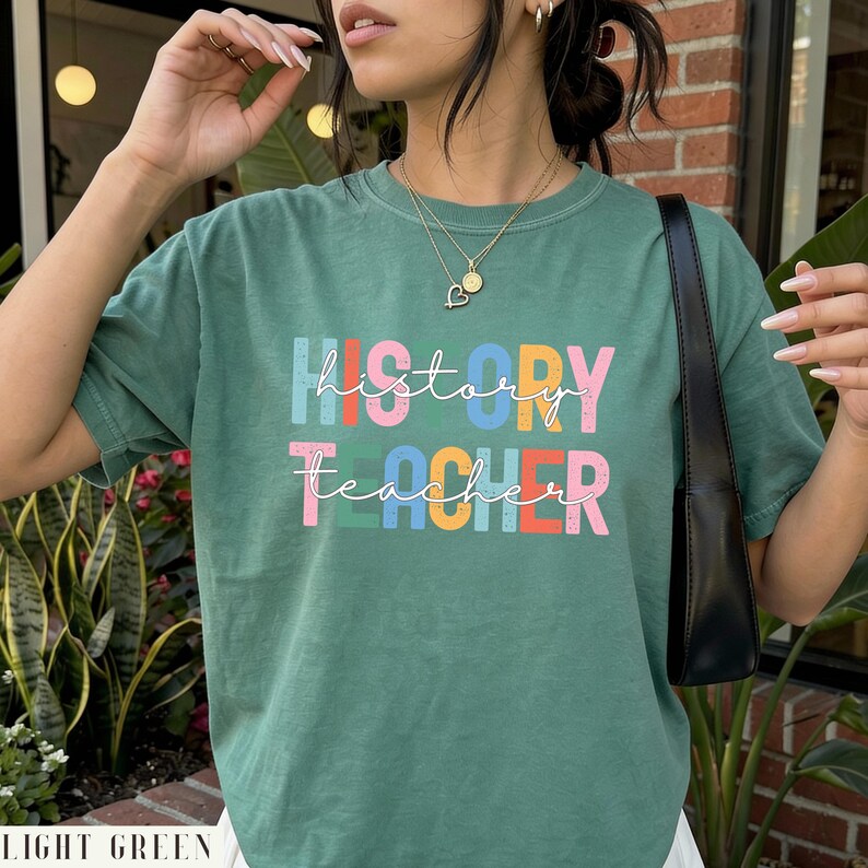 May include: A sage green short-sleeve t-shirt featuring the text "history teacher" printed in a multi-colored, distressed block font. The word "history" appears in blue, red, light blue, yellow, and pink, while "teacher" is in pink, blue, yellow, red, and pink.