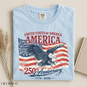 May include: Light blue t-shirt featuring a distressed American flag design with a soaring eagle and the text "AMERICA 250th Anniversary 1776-2026". The shirt is made of a soft, comfortable material.