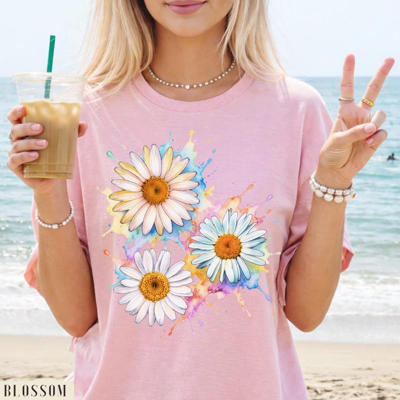 May include: A light pink t-shirt featuring a watercolor design of three daisies with white petals and orange centers. The daisies are surrounded by colorful paint splashes. The word "BLOSSOM" is printed at the bottom left.