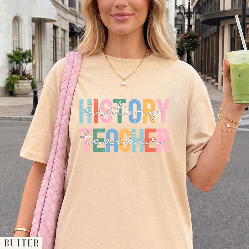 May include: A beige short-sleeved t-shirt featuring the text "HISTORY TEACHER" in a multi-colored, blocky font with a distressed texture. The word "history" is in blue, green, and yellow, while "teacher" is in green, yellow, red, and pink.