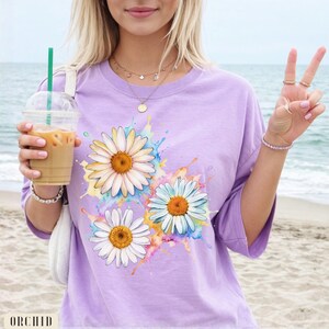 May include: A light purple t-shirt featuring a watercolor design of three daisies with white petals and yellow centers. The daisies are surrounded by colorful paint splatters. The word "ORCHID" is printed at the bottom of the shirt.