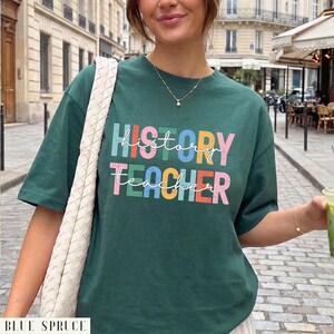 May include: A dark green short-sleeve t-shirt featuring the text "HISTORY TEACHER" in a multi-colored, distressed block font. The word "history" is printed in blue, pink, green, yellow, and orange, while "teacher" is in pink, green, yellow, orange, and blue.