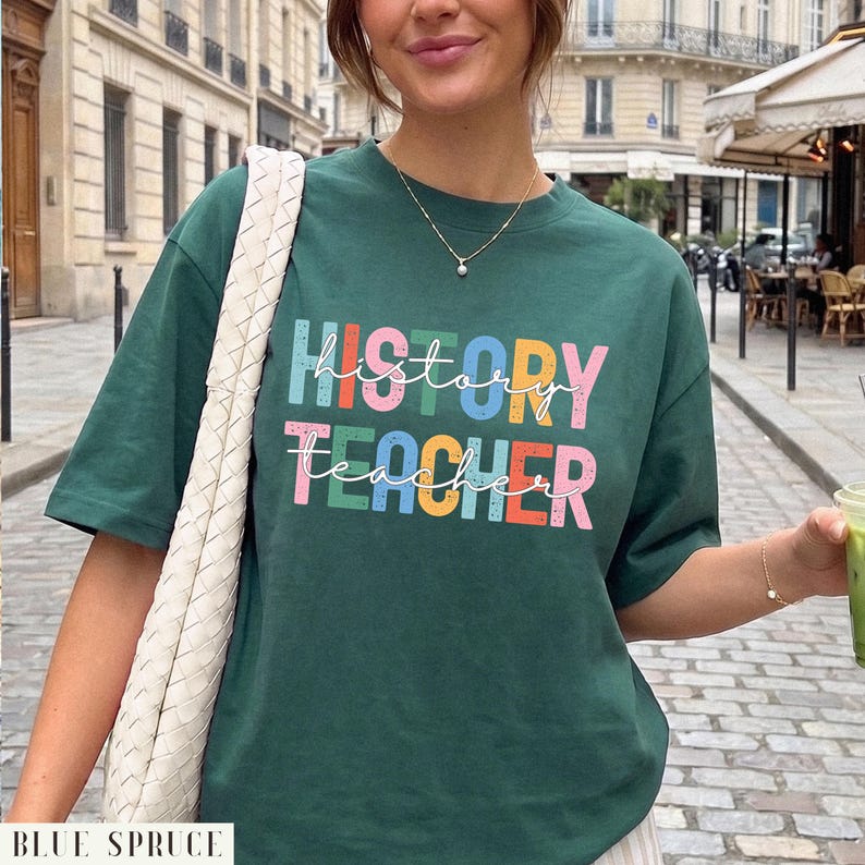 May include: A dark green short-sleeve t-shirt featuring the text "HISTORY TEACHER" in a multi-colored, distressed block font. The word "history" is printed in blue, pink, green, yellow, and orange, while "teacher" is in pink, green, yellow, orange, and blue.