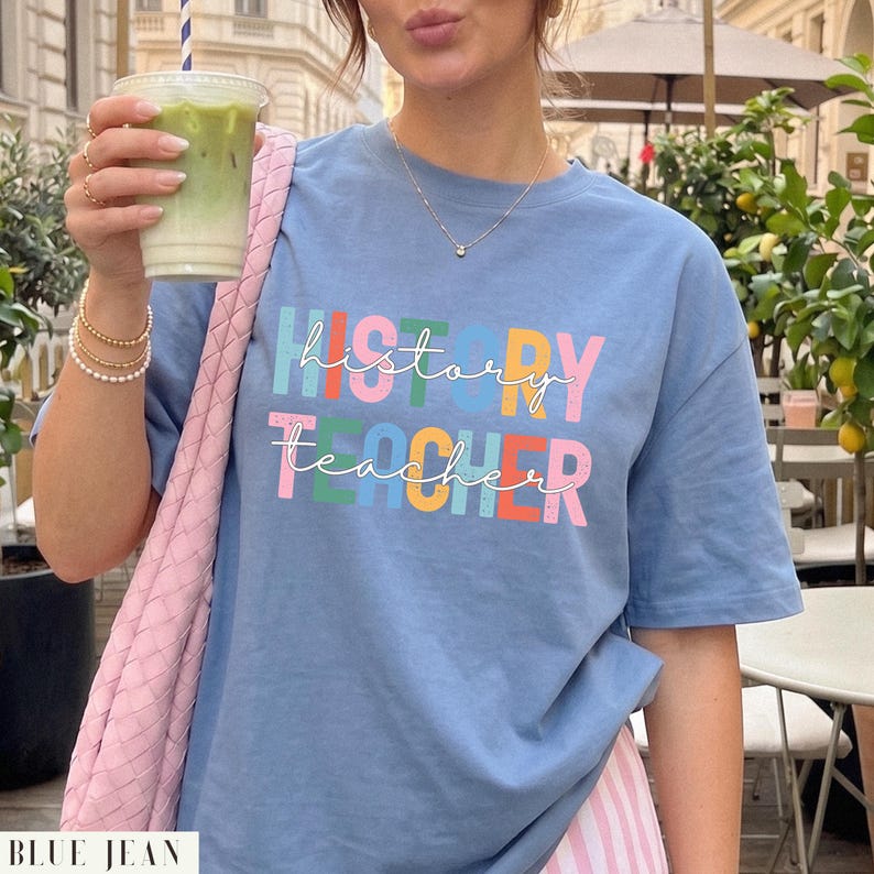 May include: A blue t-shirt featuring the text "history teacher" in a multi-colored, block-lettered font with a white script overlay. The shirt has short sleeves and a crew neckline, showcasing a casual design with a vibrant, multi-colored graphic print.