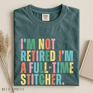 May include: Blue Spruce t-shirt with the text "I'M NOT RETIRED I'M A FULL-TIME STITCHER" in colorful letters. The shirt is folded and has a Comfort Colors tag. A book and dried plant are in the background.