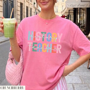 May include: A bright pink short-sleeved t-shirt featuring the text "history teacher" in white cursive script layered over block letters in various colors including teal, orange, yellow, and blue. The shirt has a crew neckline and a relaxed fit.