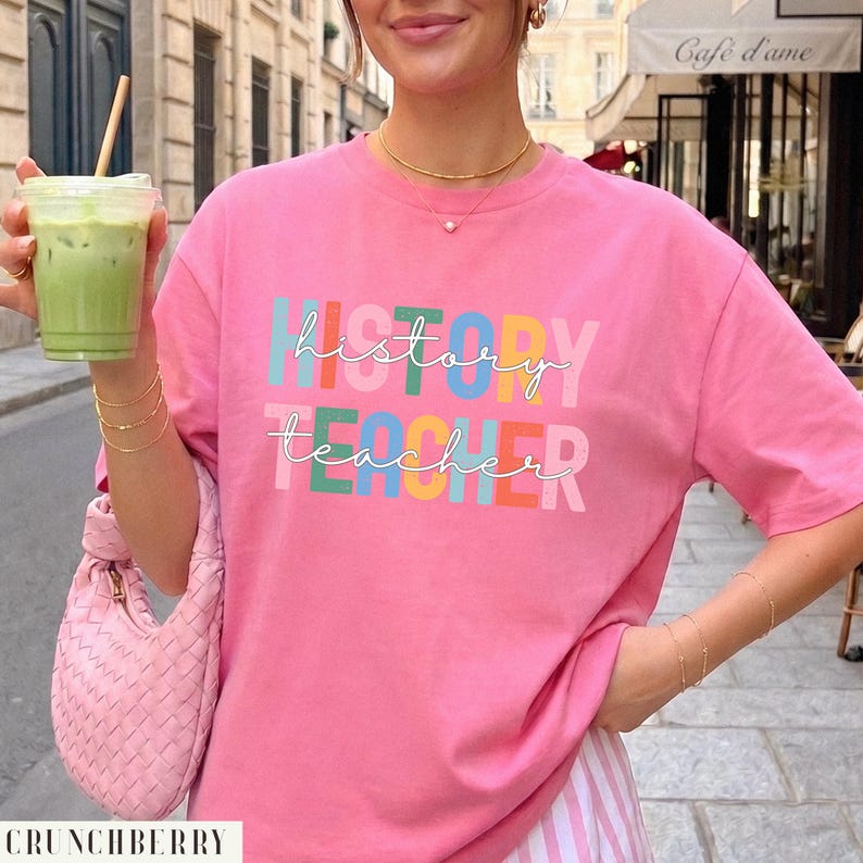 May include: A bright pink short-sleeved t-shirt featuring the text "history teacher" in white cursive script layered over block letters in various colors including teal, orange, yellow, and blue. The shirt has a crew neckline and a relaxed fit.