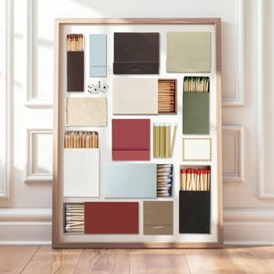 May include: A framed collection of various matchboxes in different colors, including brown, blue, red, and green. Some boxes are open, revealing the matches inside. Two dice are also included in the arrangement. The matches have red or green tips.