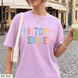 May include: A light purple t-shirt featuring the text HISTORY TEACHER in a multi-colored block font with white cursive script layered over it. The shirt has short sleeves and a crew neckline, paired with a white shoulder bag and gold jewelry.