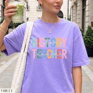 May include: A light purple short-sleeve t-shirt featuring the text "history teacher" in a multi-colored block font. The design includes stacked letters in shades of green, blue, yellow, orange, and pink, with white cursive script overlaying the text.