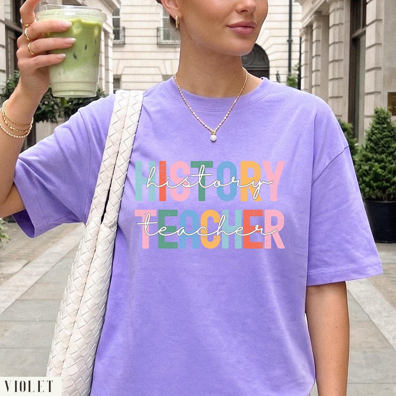 May include: A light purple short-sleeve t-shirt featuring the text "history teacher" in a multi-colored block font. The design includes stacked letters in shades of green, blue, yellow, orange, and pink, with white cursive script overlaying the text.