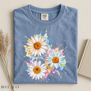 May include: A blue t-shirt with a watercolor design of three daisies. The daisies have white petals and yellow centers, with colorful paint splashes in the background. The shirt has a tag that says "Comfort Colors".