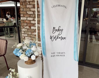 Könnte beinhalten: Ein weißes Stoffbanner auf einem rechteckigen Rahmen mit dem schwarzen Schriftzug "CELEBRATING", "Baby Workman" und "SAM + SARAH'S BABY SHOWER". Das Banner ist mit zwei langen, hellblauen Bändern verziert, die an den oberen Ecken befestigt sind.
