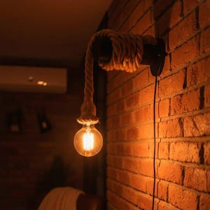 Może przedstawiać: Rustykalna lampa ścienna z żarówką Edison w stylu vintage. Oprawa posiada konstrukcję ze zwiniętej liny i czarną metalową podstawę, zamontowaną na czerwonej ceglanej ścianie. Ciepłe światło żarówki tworzy przytulną atmosferę. Światło jest połączone z czarnym przewodem.
