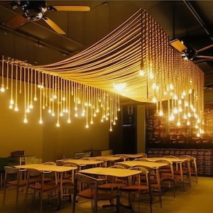 Puede incluir: Interior de un restaurante con una iluminación única. Numerosas bombillas cuelgan de cuerdas, creando un brillo cálido y amarillo. El comedor cuenta con mesas y sillas de madera. También se ven ventiladores de techo.