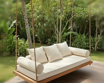 May include: A rectangular wooden hanging daybed suspended by thick ropes, featuring a flat base, a long off-white mattress, three square off-white throw pillows, and two cylindrical off-white bolster pillows positioned at each end of the seat.