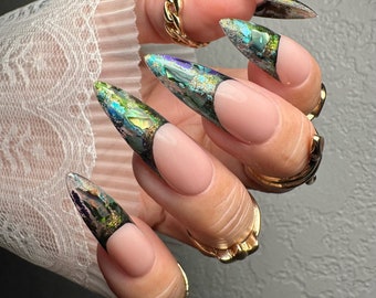May include: Close-up of long, almond-shaped nails with a French manicure. The tips are adorned with iridescent, multi-colored foil, creating a shimmering effect. Gold rings accent the fingers, adding a touch of elegance to the nail art.
