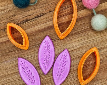 May include: Three orange leaf-shaped cookie cutters and three purple leaf-shaped clay pieces with an embossed fern-like pattern. The orange cutters have a hollow center, while the purple pieces are solid with a textured surface detail.