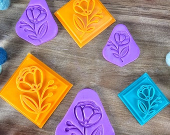 May include: A collection of six plastic clay cutters in various shapes and colors, including orange diamond shapes, purple shield shapes, and a blue diamond shape, each featuring a raised floral design with a stem and leaves.