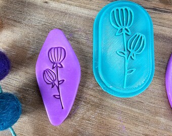 May include: A purple diamond-shaped clay cutter and a teal oval-shaped clay cutter, both featuring a raised line design of two tulip-like flowers on stems. The cutters have a smooth, matte finish and are positioned on a wooden surface.