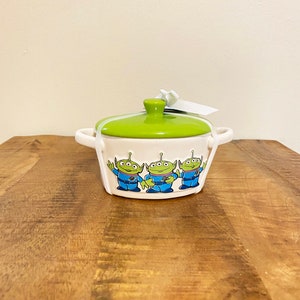 May include: A white ceramic soup pot with a green lid and handles. The pot features a design of four green aliens from the Toy Story movie.