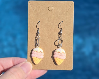 May include: A pair of silver dangle earrings with a pink and yellow ice cream cone charm. The earrings are displayed on a brown card.