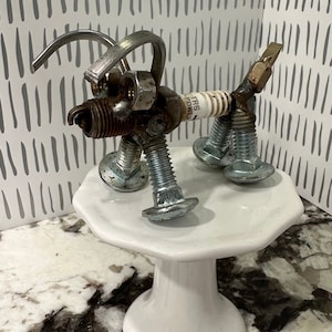 May include: A metal sculpture of a dog made from nuts, bolts, and a spark plug. The dog is standing on a white cake stand. The spark plug has the text "TRS POWER" on it.