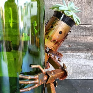 May include: A whimsical, handcrafted metal sculpture of a robot holding a small succulent planter. The robot is bronze-colored with a distressed finish, and the succulent has green leaves. The sculpture is positioned next to a large green glass bottle.