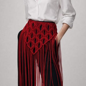 Puede incluir: Una mujer con una camisa blanca y pantalones negros. Un cinturón de macramé rojo con un patrón geométrico y largas franjas es el punto focal del atuendo. El cinturón añade un toque bohemio único al conjunto.
